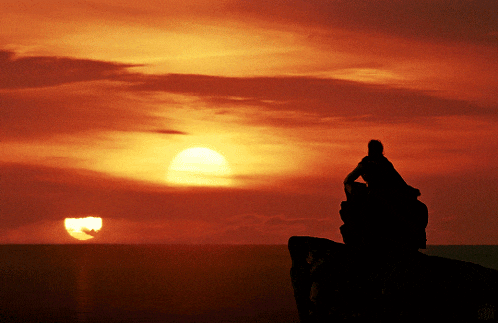 a person sits on a cliff watching the sun set over the ocean
