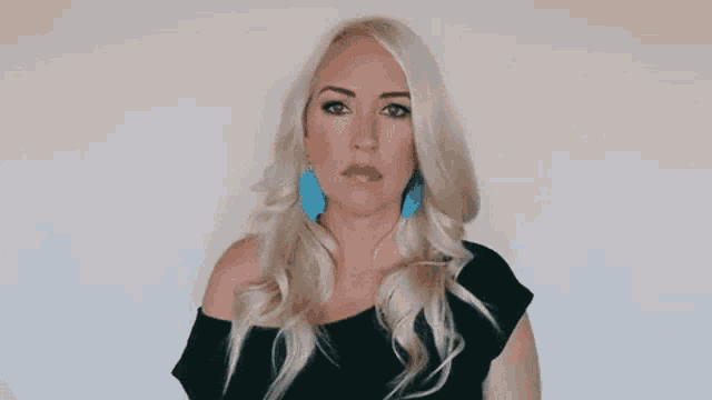 a woman with blonde hair and blue earrings is wearing a black top and making a funny face .