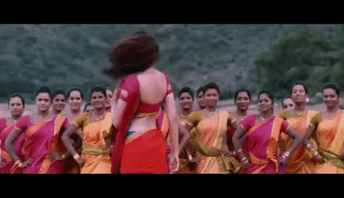 a woman in a red saree is dancing in front of a large group of women in pink and yellow dresses .