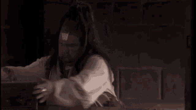 a man with long hair is sitting at a table in a dark room holding something .