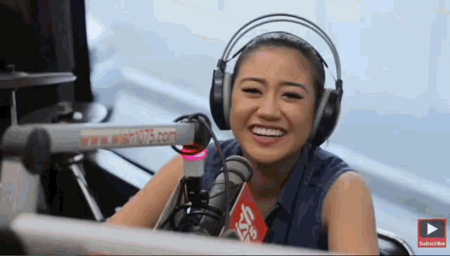 a woman is wearing headphones and smiling in front of a microphone with a subscribe button in the corner