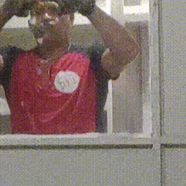 a man wearing a red shirt with a white circle on the shoulder is looking out a window