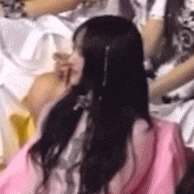 a woman with long black hair is wearing a pink dress and sitting in a crowd of people .