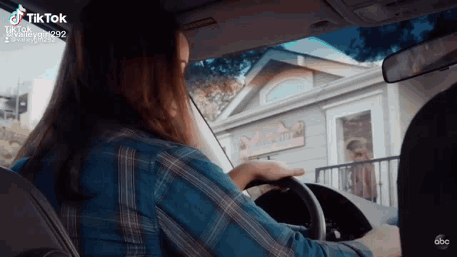 a woman in a blue plaid shirt is driving a car with a house in the background .