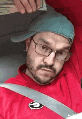 a man with a beard and glasses is wearing a hat and a seat belt while sitting in a car .