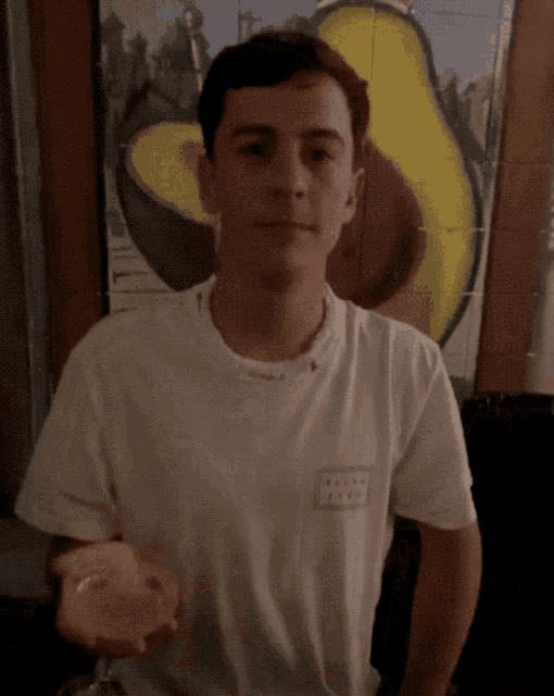 a young man wearing a white t-shirt with the word avocado on the back is standing in front of an avocado painting
