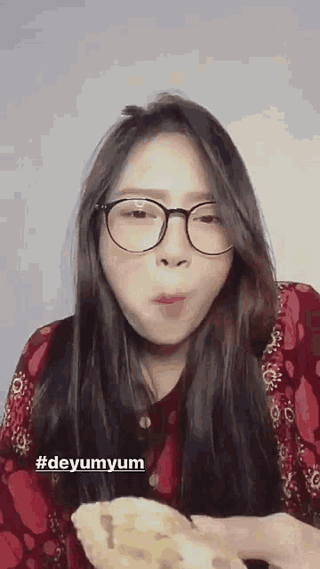 a woman wearing glasses and a red shirt is eating a sandwich .