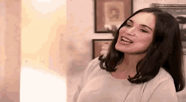 a woman in a white shirt is smiling and looking at the camera while standing in a living room .
