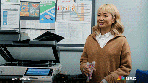 a woman in a brown sweater stands in front of a printer with a nbc logo on it