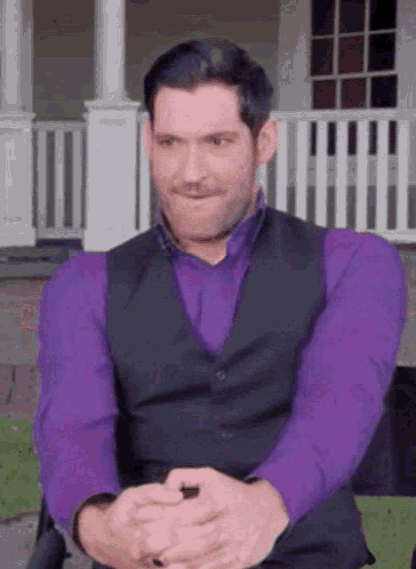 a man in a purple shirt and a black vest is sitting in front of a house .