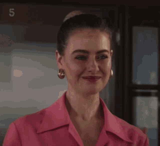 a woman in a pink shirt and gold earrings smiles in front of a sign that says 5