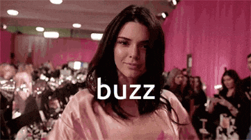 a woman in a pink shirt is standing in front of a pink curtain with the word buzz written on it .