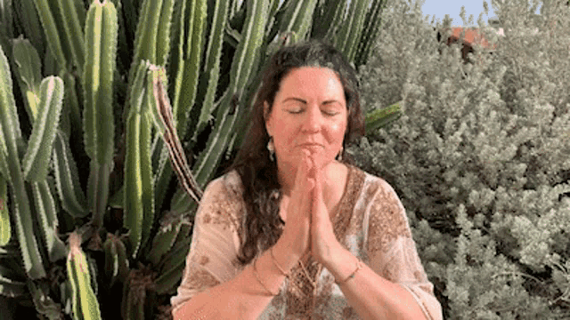 a woman is sitting in front of a cactus with her eyes closed and her hands folded in prayer .