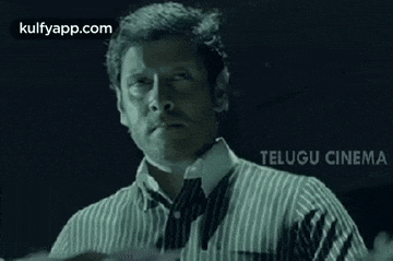 a man in a striped shirt and tie is standing in front of a telugu cinema sign .