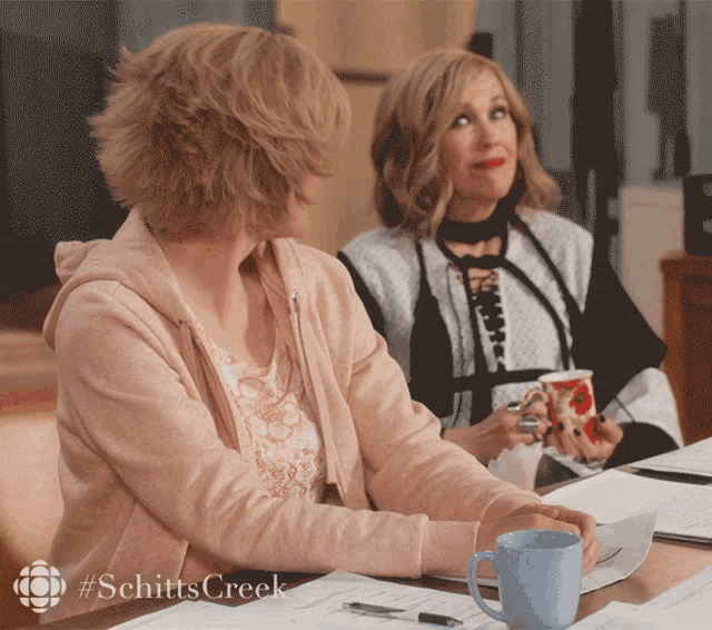 two women sit at a table with #schittscreek written on the bottom right
