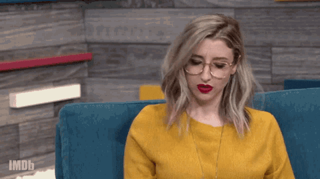 a woman wearing glasses and a yellow sweater is sitting on a blue couch with imdb written on the bottom