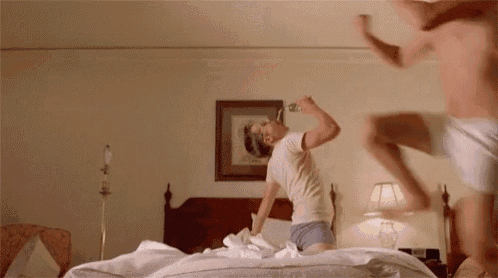 a man is drinking from a bottle while another man jumps on a bed .