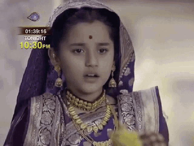 a little girl is wearing a purple dress and gold jewelry and holding a pear .