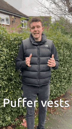 a man wearing a black puffer vest is standing in front of a hedge .