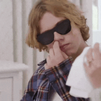 a young man wearing sunglasses and a plaid shirt is holding a napkin to his nose .