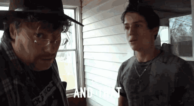 a man wearing a hat and glasses stands next to another man who is wearing a shirt that says and that