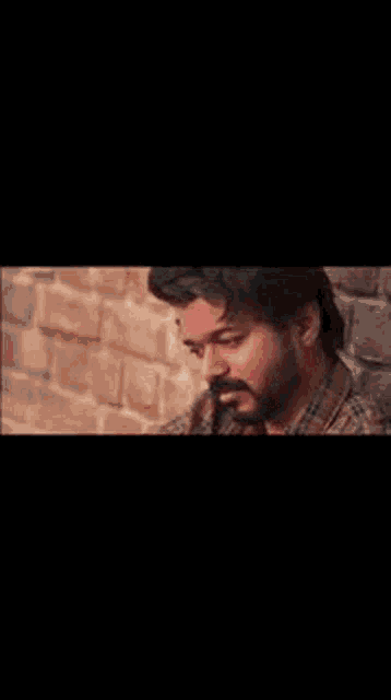 a man with a beard is standing in front of a brick wall and looking down .