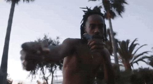 a shirtless man with dreadlocks is smoking a cigarette in front of a palm tree .