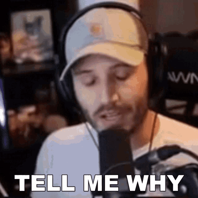 a man wearing headphones and a hat is standing in front of a microphone and saying `` tell me why '' .