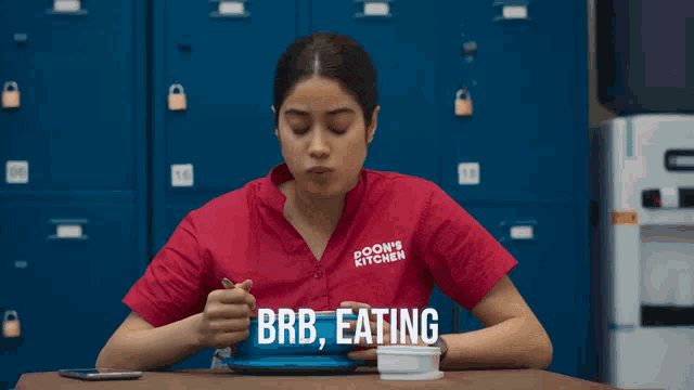 a woman wearing a red shirt that says boon 's kitchen is eating a bowl of food