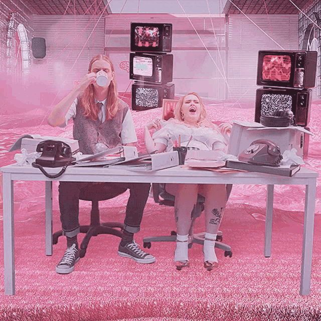a man drinking from a cup while a woman sits at a desk in front of a stack of televisions