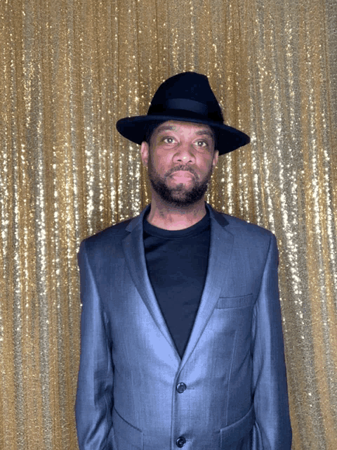 a man wearing a black hat and a grey suit stands in front of a gold sequined backdrop