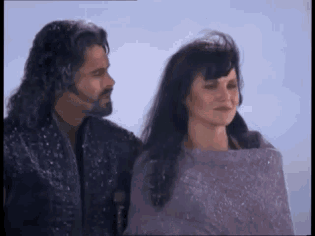 a man and a woman are standing in the snow . the woman is wrapped in a purple blanket .