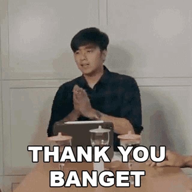 a man is sitting at a table with his hands folded and the words thank you banget written on it .