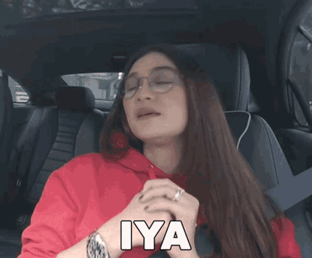 a woman wearing glasses and a red sweatshirt is sitting in a car with the word iya written on her face