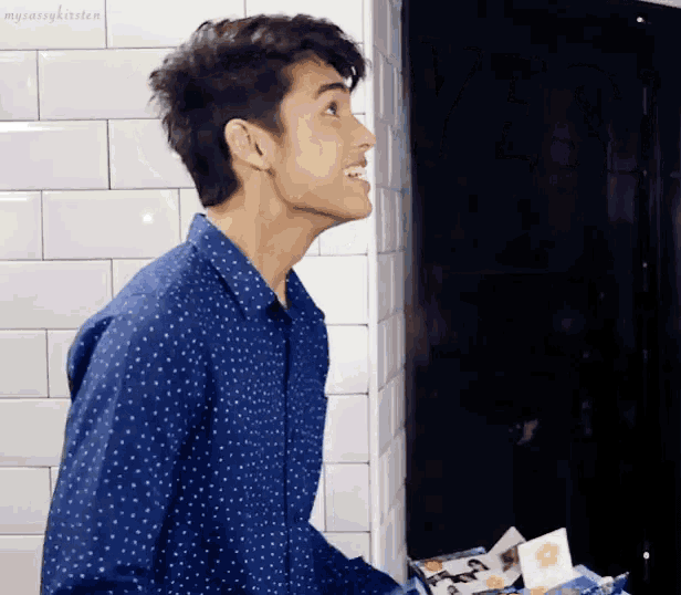 a man in a blue polka dot shirt stands in front of a white tile wall