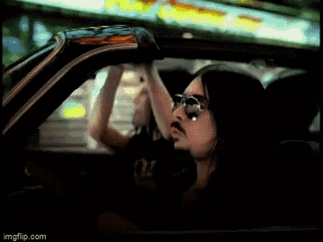a man with long hair and glasses is driving a car