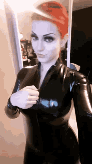 a woman with red hair is wearing a black latex suit and taking a selfie in front of a mirror .
