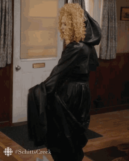 a woman in a black cape is standing in front of a door with #schittscreek on the bottom