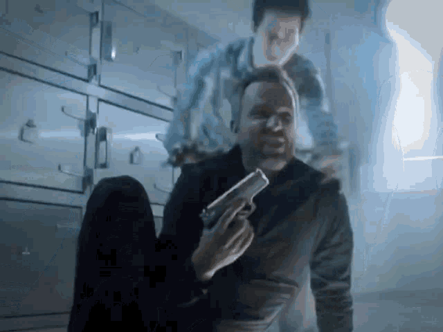 a man is sitting on the floor holding a gun while another man is standing behind him .