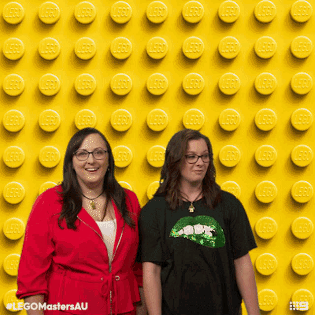 two women standing in front of a wall of yellow lego blocks