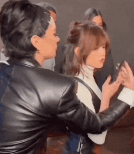 a woman in a leather jacket is taking a picture of another woman in a white turtleneck .
