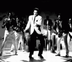 a black and white photo of elvis presley dancing on stage with a band behind him .