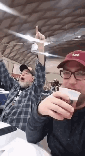 two men are sitting at a table with their arms in the air and one is wearing a hat that says albe