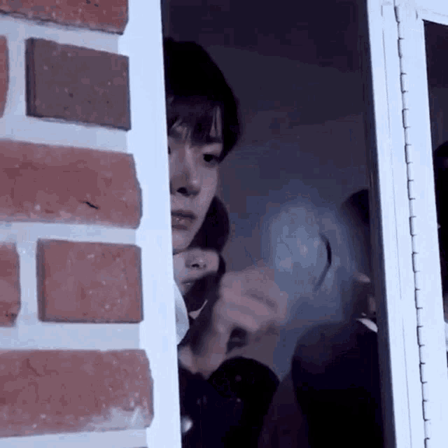 a man is peeking out of a window next to a brick wall while a woman looks on .