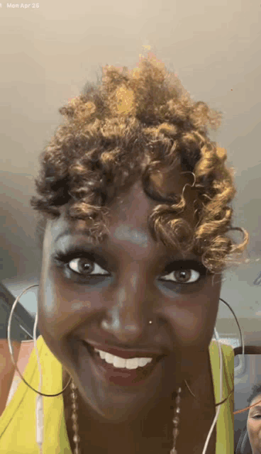 a woman wearing hoop earrings and a yellow tank top smiles for the camera on monday april 15