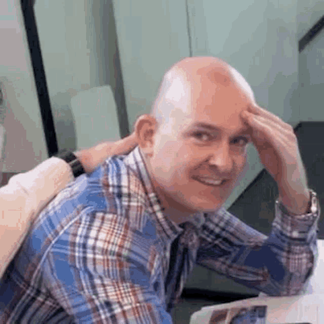 a bald man in a plaid shirt is sitting at a table with his hands on his head .
