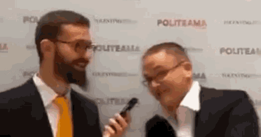 a man in a suit and tie is talking into a microphone while another man looks on ..