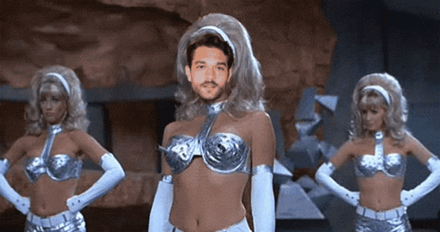 a man in a wig is standing in front of two women in silver outfits