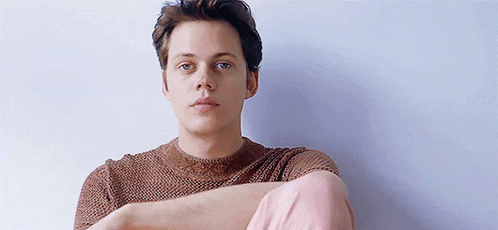 a young man wearing a brown sweater and pink pants is sitting with his arms crossed and looking at the camera .