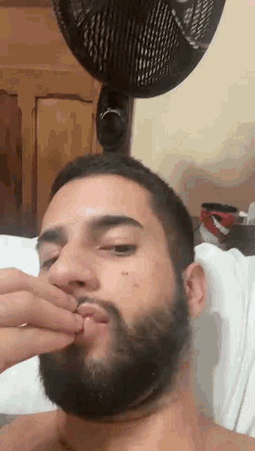 a man with a beard is laying in bed with a fan behind him